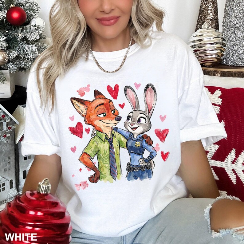 Może przedstawiać: Biała koszulka z kresk&oacute;wkową ilustracją Nicka Wilde'a i Judy Hopps z filmu Zwierzogr&oacute;d, otoczona czerwonymi sercami. Nick ma na sobie zieloną koszulę, a Judy mundur policjantki. Słowo "WHITE" jest widoczne na dole obrazu.