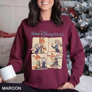 Puede incluir: Sudadera granate con un dise&ntilde;o gr&aacute;fico de cuatro paneles de personajes de Zootopia. El panel superior tiene el texto "Walt Disney World". La parte inferior de la sudadera tiene la palabra "MAROON".