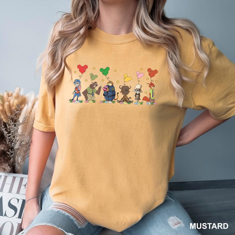 May include: Mustard-colored t-shirt featuring a whimsical cartoon design of several animal characters holding heart-shaped balloons. The design includes a variety of colors and is printed on a comfortable, casual tee.