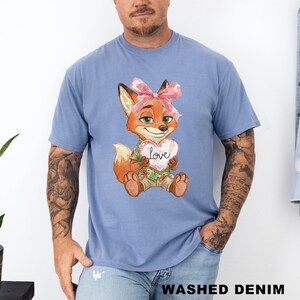 May include: A light blue t-shirt featuring a cartoon fox holding a heart-shaped sign with the word "Love". The fox has orange fur, green eyes, and a pink bow. The shirt is made of a soft material.