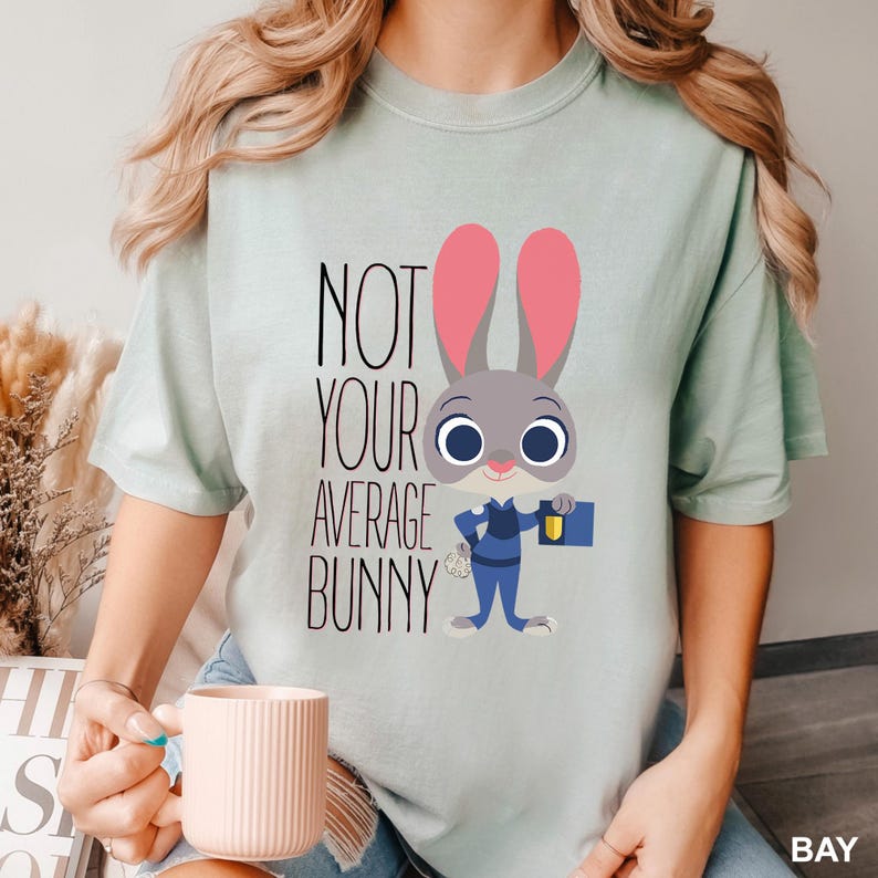 May include: A pale green t-shirt with a cartoon bunny in a police uniform. The text "NOT YOUR AVERAGE BUNNY" is printed in black. The bunny has pink ears and is holding a blue folder. The shirt is a solid colour.