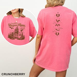 May include: A pink t-shirt with a graphic on the front featuring a castle, trees, and the text "Riorrson House". The back of the shirt has a vertical design with dragons and clouds. The shirt is a solid color and has short sleeves.