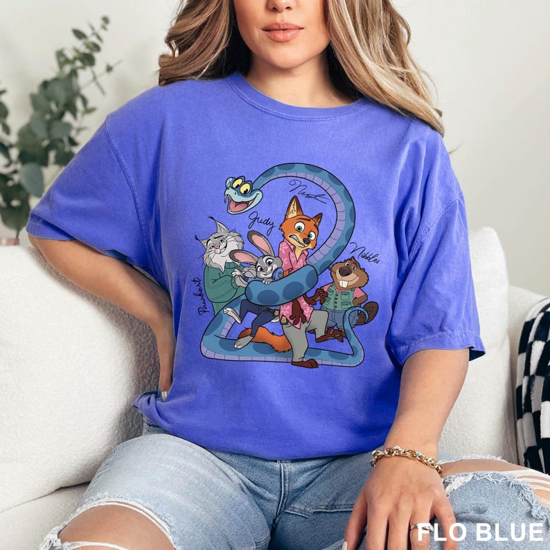 May include: A blue t-shirt featuring a cartoon graphic of characters from a movie. The graphic includes a fox, a rabbit, a beaver, and a cat, all wrapped in a blue snake. The shirt has the text "FLO BLUE" at the bottom.