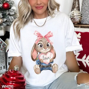 May include: White t-shirt featuring a cartoon bunny holding a heart with the word "Love". The bunny has a pink bow, and is wearing a blue uniform. The shirt is a casual, oversized fit.