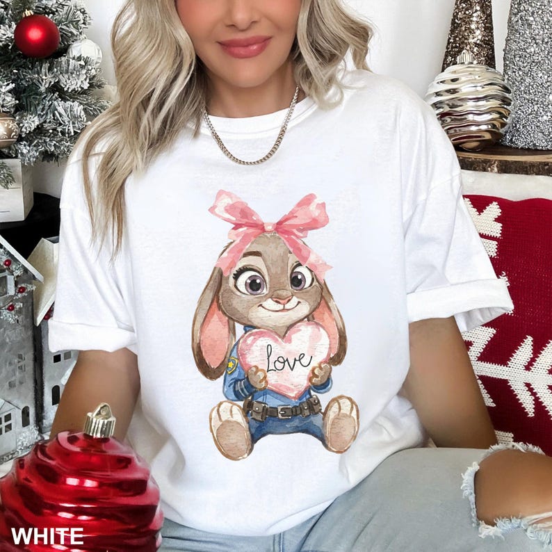 May include: White t-shirt featuring a cartoon bunny holding a heart with the word "Love". The bunny has a pink bow, and is wearing a blue uniform. The shirt is a casual, oversized fit.