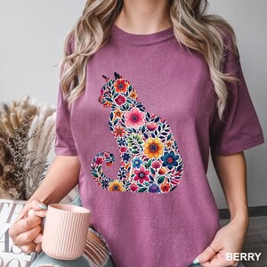 May include: Berry-colored t-shirt featuring a floral cat design. The cat silhouette is filled with colorful flowers and leaves in shades of pink, blue, yellow, and green. The word "BERRY" is printed in white at the bottom right.