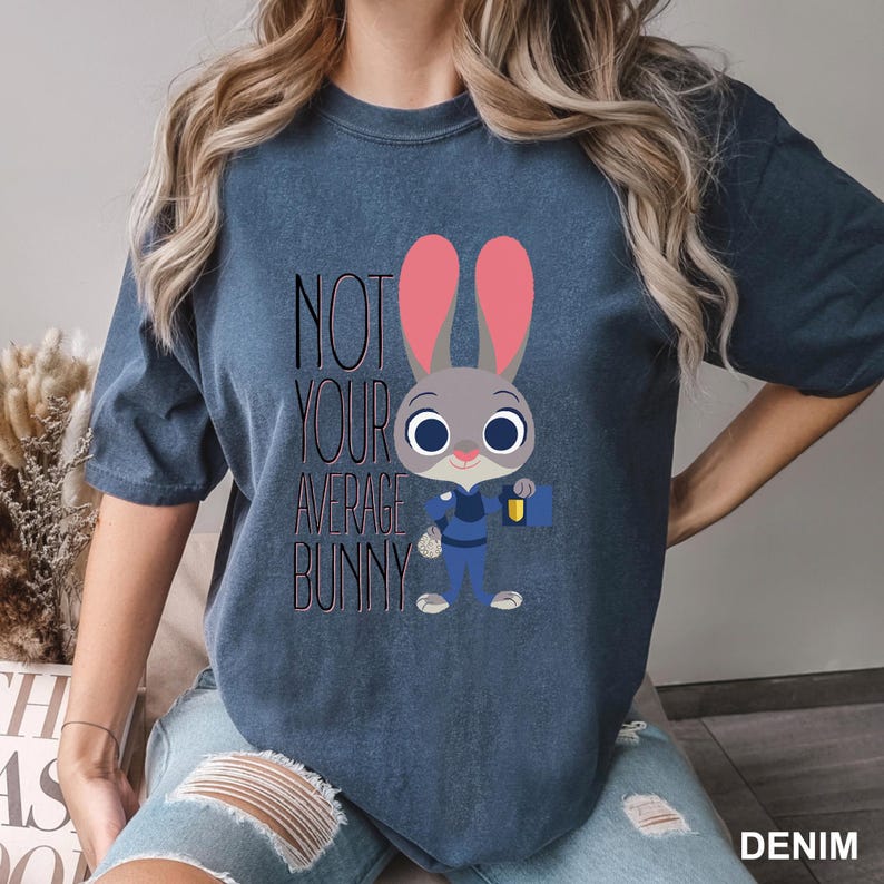 May include: Denim blue t-shirt with a cartoon bunny character in a police uniform and pink ears. The text "NOT YOUR AVERAGE BUNNY" is printed above the character. The shirt is a casual style.