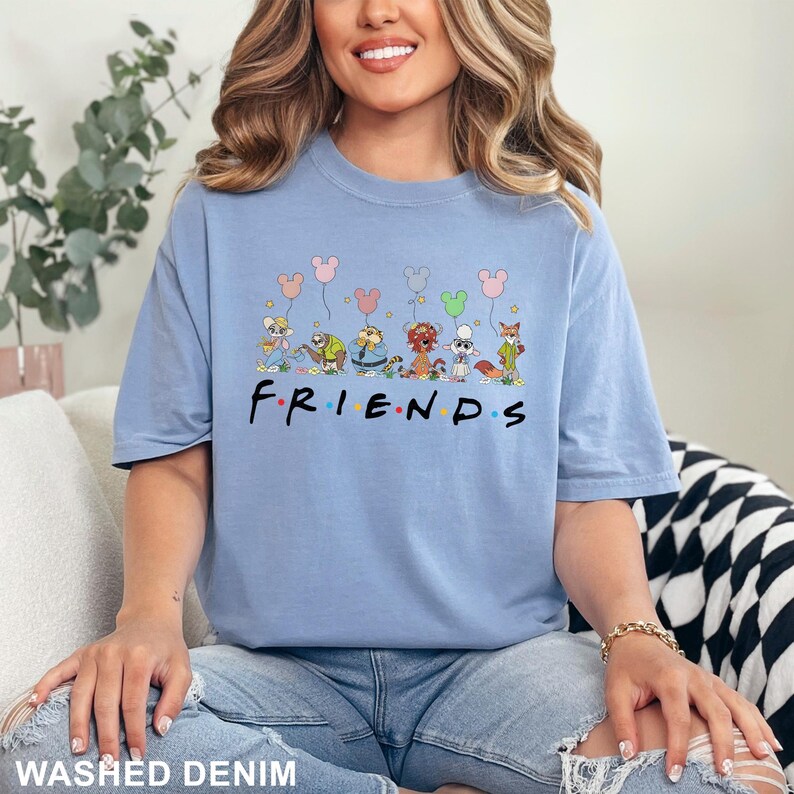 May include: A light blue t-shirt with the word "FRIENDS" in black and colorful letters. Above the word are cartoon characters holding balloons. The shirt is made of a soft material and is a washed denim color.