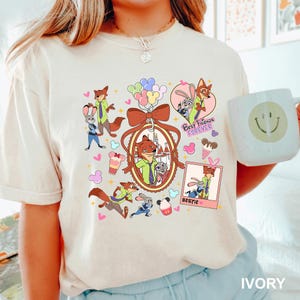 Op de afbeelding: Ivoor kleurig T-shirt met stripfiguren, waaronder een vos en een konijn, met de woorden "Best Friends Forever" en "Bestie". Het ontwerp bevat harten, ijsjes en ballonnen. Het shirt heeft een comfortabele pasvorm.