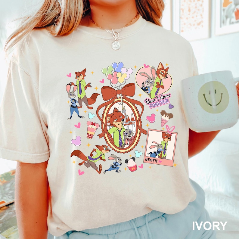 Op de afbeelding: Ivoor kleurig T-shirt met stripfiguren, waaronder een vos en een konijn, met de woorden "Best Friends Forever" en "Bestie". Het ontwerp bevat harten, ijsjes en ballonnen. Het shirt heeft een comfortabele pasvorm.