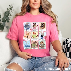 Op de afbeelding: Een felroze T-shirt met korte mouwen en een raster van negen kaders in polaroidstijl. Elk kader bevat een Zootopia-personage met de naam eronder gedrukt, waaronder Chief Bogo, Judith, Mr. Big, Finnick, Nick Wilde, Judy Hopps, Gary de'snack, Flash en Nibbles Maplestick.
