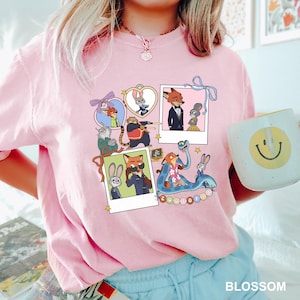 May include: Pale pink crew neck t-shirt with a vibrant graphic of Zootopia characters. The design incorporates polaroid-style images of the characters, with the word "Zootopia" at the bottom. A white mug with a smiley face is visible.