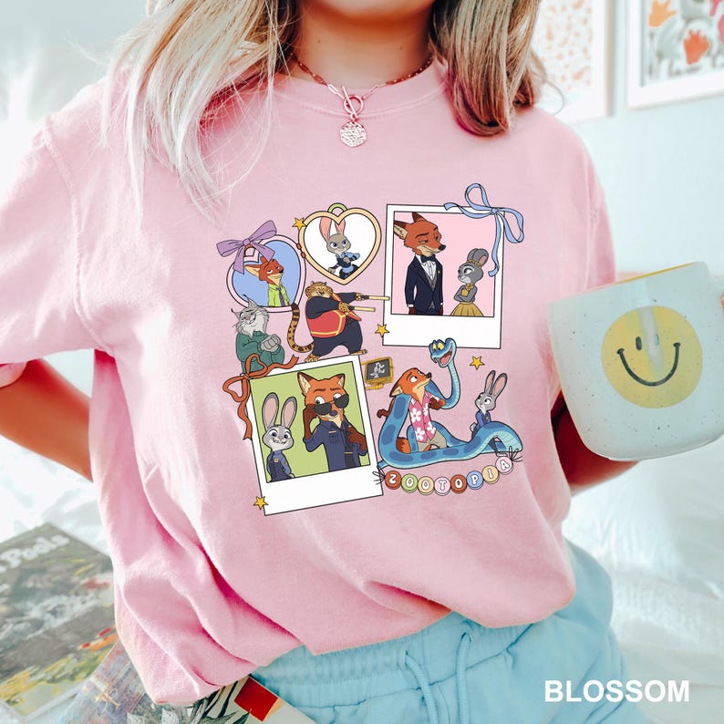 May include: Pale pink crew neck t-shirt with a vibrant graphic of Zootopia characters. The design incorporates polaroid-style images of the characters, with the word "Zootopia" at the bottom. A white mug with a smiley face is visible.