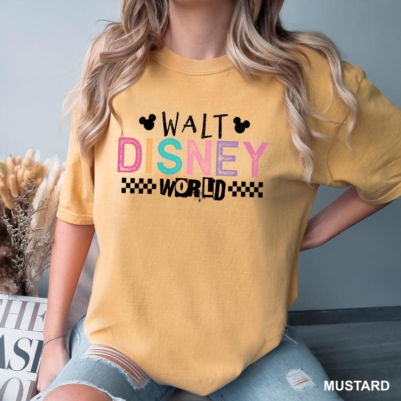 May include: A mustard yellow short-sleeve t-shirt featuring the text WALT DISNEY WORLD in a multi-colored, distressed font. The design includes two small black Mickey Mouse silhouettes and black checkered patterns flanking the word WORLD.