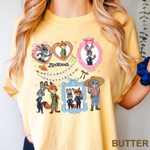 Op de afbeelding: Boterkleurig T-shirt met Zootopia-personages. Het ontwerp bevat ingelijste afbeeldingen van Judy Hopps en Nick Wilde, het woord "Zootopia" en de zin "Welcome to Zootopia". Het shirt toont verschillende illustraties van de personages.