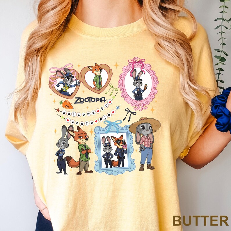 Op de afbeelding: Boterkleurig T-shirt met Zootopia-personages. Het ontwerp bevat ingelijste afbeeldingen van Judy Hopps en Nick Wilde, het woord "Zootopia" en de zin "Welcome to Zootopia". Het shirt toont verschillende illustraties van de personages.