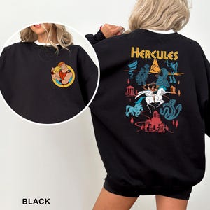 K&ouml;nnte beinhalten: Schwarzes Sweatshirt mit einem Cartoon-Motiv von Herkules auf der Vorderseite und einem gr&ouml;&szlig;eren Design auf der R&uuml;ckseite. Das R&uuml;ckendesign enth&auml;lt das Wort "Hercules" in Gold sowie verschiedene Zeichentrickfiguren und Szenen aus dem Film. Das Sweatshirt hat einen wei&szlig;en Kragen.