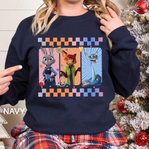 Puede incluir: Sudadera azul marino con un dise&ntilde;o de dibujos animados de tres personajes en marcos rectangulares. Los personajes son un conejo con uniforme de polic&iacute;a, un zorro con camisa y corbata verdes y una serpiente azul. La palabra "NAVY" est&aacute; impresa en la parte inferior izquierda.