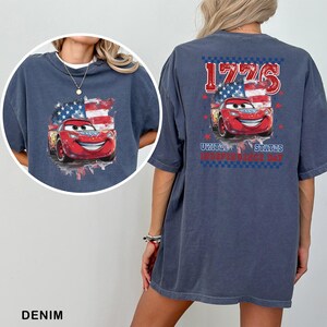 May include: A dark blue t-shirt featuring a graphic of a red race car with a smiling face, positioned in front of a stylised American flag. The back design includes the text 1776 and United States Independence Day in red and blue.