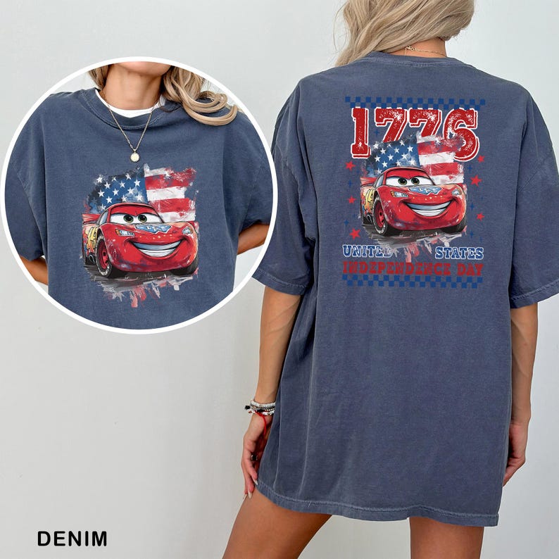 May include: A dark blue t-shirt featuring a graphic of a red race car with a smiling face, positioned in front of a stylised American flag. The back design includes the text 1776 and United States Independence Day in red and blue.