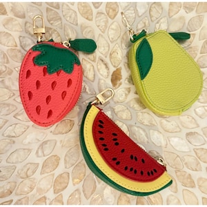 May include: Three fruit-shaped coin purses: a red strawberry, a yellow-green pear, and a watermelon slice. Each purse has a gold-colored zipper and a small loop for attaching to a keychain. The strawberry has a green stem and leaf.