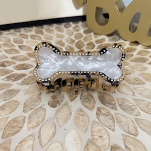 May include: A bone-shaped hair claw clip with a tortoiseshell pattern and clear rhinestones. The center of the clip is white and marbled. The clip is made of plastic and has a secure grip.