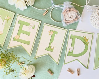 May include: Green and white banner with the word "EID" in large, green letters. The letters are decorated with small white flowers and green leaves. The banner is strung with twine and small wooden clothespins.