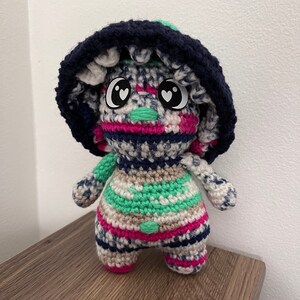 Può includere: Una figura di fungo all'uncinetto fatta a mano con un cappello blu navy e un corpo a righe nei toni del rosa, verde, bianco e blu. Il fungo ha grandi occhi a forma di cuore e un'espressione carina. La figura è su una superficie di legno.