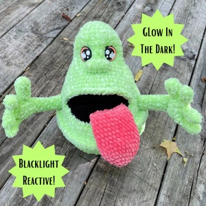 Può includere: Un giocattolo di peluche verde lime con una bocca spalancata e una lingua rosa. Il giocattolo ha grandi occhi da cartone animato e braccia tese. Il testo sull'immagine recita "Glow in the Dark!" e "Blacklight Reactive!"