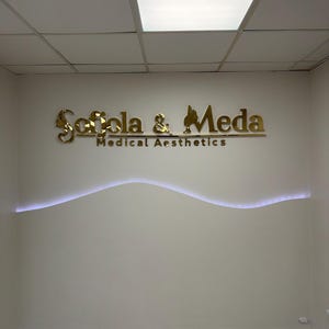 May include: A wall-mounted sign featuring gold-colored, reflective lettering that reads Sofola & Meda Medical Aesthetics. The sign is positioned above a horizontal, wavy, white LED light strip integrated into the beige wall surface.