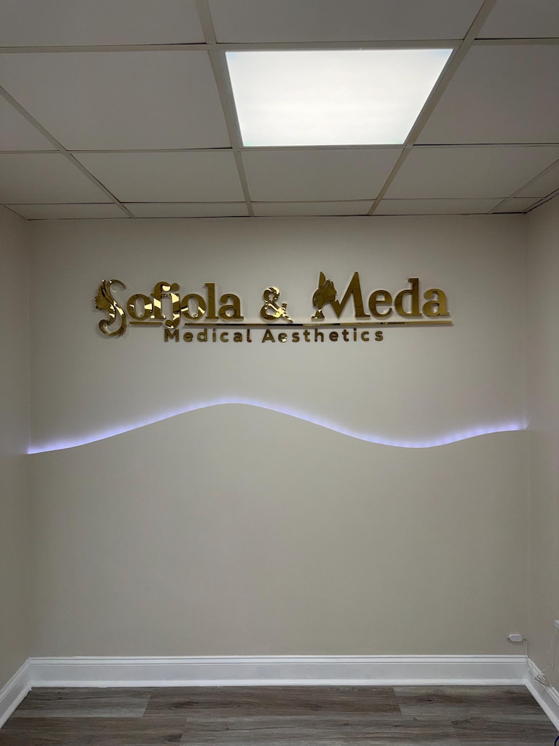 May include: A wall-mounted sign featuring gold-colored, reflective lettering that reads Sofola & Meda Medical Aesthetics. The sign is positioned above a horizontal, wavy, white LED light strip integrated into the beige wall surface.