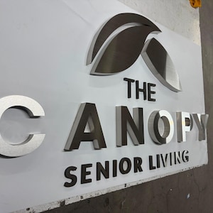 May include: A three-dimensional wall sign featuring the text THE CANOPY SENIOR LIVING in raised, metallic silver and dark grey letters. Above the text is a stylized leaf icon in a brushed silver finish, all mounted on a flat white panel.