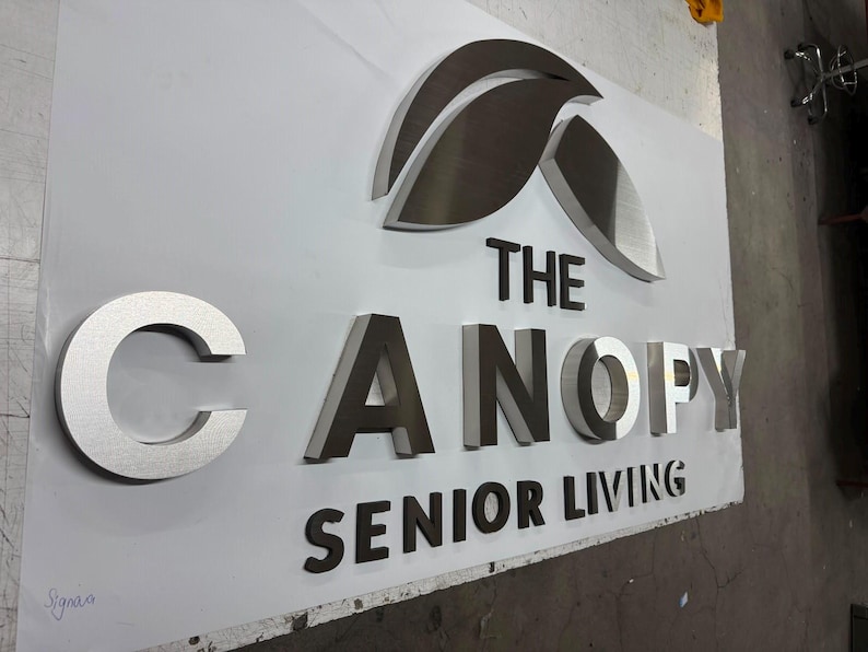 May include: A three-dimensional wall sign featuring the text THE CANOPY SENIOR LIVING in raised, metallic silver and dark grey letters. Above the text is a stylized leaf icon in a brushed silver finish, all mounted on a flat white panel.