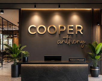 Könnte beinhalten: Moderner Büroeingangsbereich mit einer dunkelgrauen Wand mit goldenen Buchstaben "COOPER" über dem Namen "anthony" in Schreibschrift. Ein schwarzer Empfangstresen mit einem Schild "BREAKTHROUGH THERAPEUTIC CONCEPTS" befindet sich darunter. Topfpflanzen flankieren den Schreibtisch.