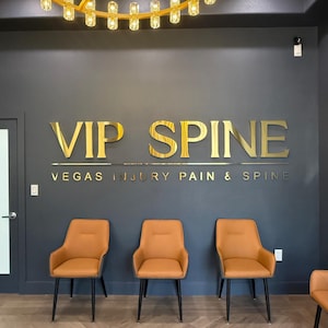 May include: A dark gray wall features gold metallic lettering that reads VIP SPINE and VEGAS INJURY PAIN & SPINE. Three orange chairs with black legs are arranged in a row against the wall, beneath a gold circular chandelier with glass lights.