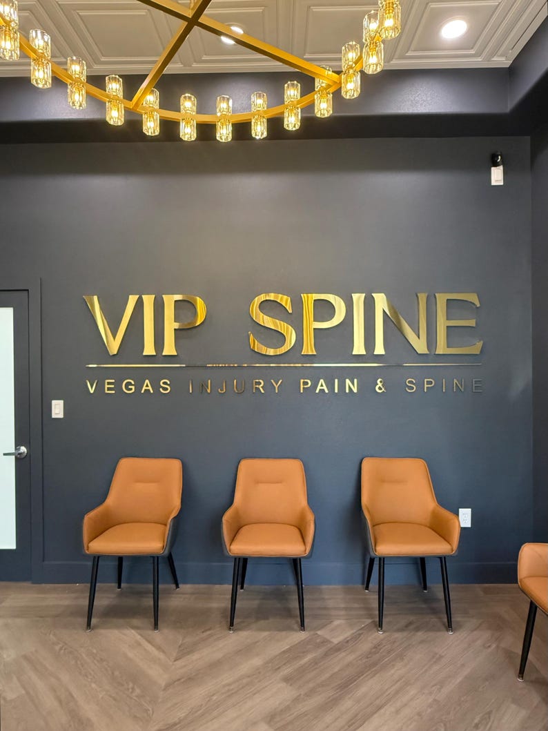 May include: A dark gray wall features gold metallic lettering that reads VIP SPINE and VEGAS INJURY PAIN & SPINE. Three orange chairs with black legs are arranged in a row against the wall, beneath a gold circular chandelier with glass lights.