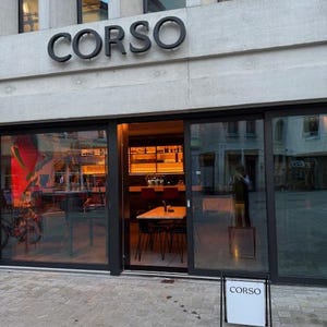 May include: The storefront of a business named CORSO features large glass windows and a sliding door entrance. Above the entrance, the word CORSO is displayed in large black letters. A white sandwich board sign with the word CORSO sits outside.