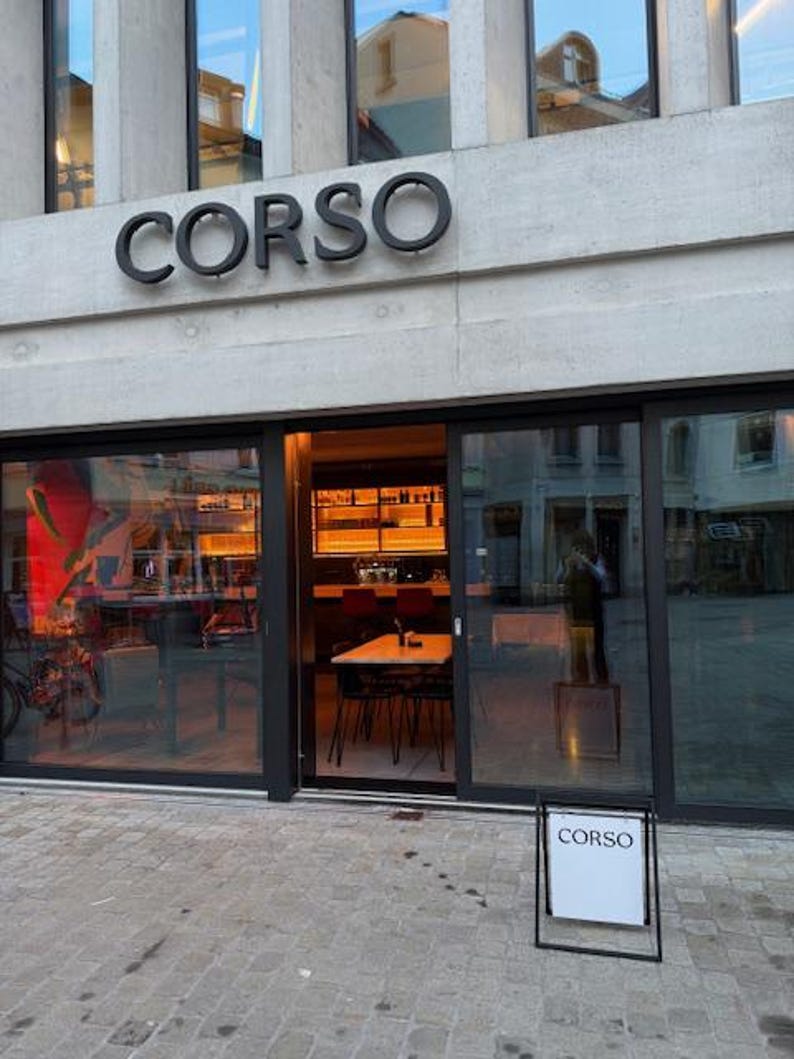 May include: The storefront of a business named CORSO features large glass windows and a sliding door entrance. Above the entrance, the word CORSO is displayed in large black letters. A white sandwich board sign with the word CORSO sits outside.