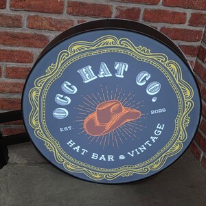 May include: Round, vintage-style sign for "OGG HAT CO." with a cowboy hat illustration. The sign is dark blue with gold and white accents, and the text "HAT BAR & VINTAGE" is visible. The sign is mounted on a black bracket against a brick wall.