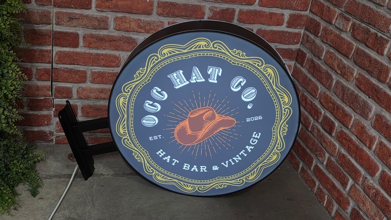 May include: Round, vintage-style sign for "OGG HAT CO." with a cowboy hat illustration. The sign is dark blue with gold and white accents, and the text "HAT BAR & VINTAGE" is visible. The sign is mounted on a black bracket against a brick wall.