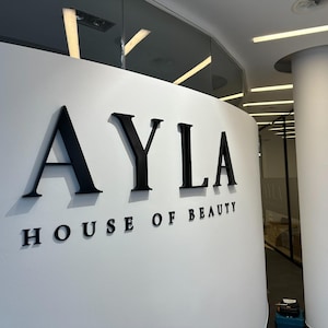 May include: A white curved wall featuring the black three-dimensional lettering AYLA HOUSE OF BEAUTY. The letters are mounted on the surface, creating a clean and professional display for the business signage in a bright, modern interior space.