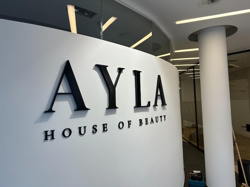 May include: A white curved wall featuring the black three-dimensional lettering AYLA HOUSE OF BEAUTY. The letters are mounted on the surface, creating a clean and professional display for the business signage in a bright, modern interior space.