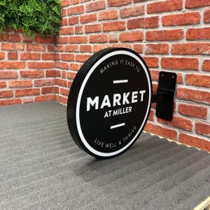 Pode incluir: Uma placa redonda em preto e branco com o texto "MARKET AT MILLER" está montada em uma parede de tijolos. A placa também inclui as frases "MAKING IT EASY TO" e "LIVE WELL & THRIVE". Um suporte de metal preto suporta a placa.