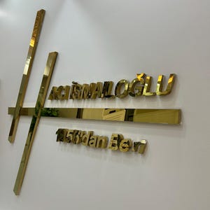 Może przedstawiać: Złoty, tr&oacute;jwymiarowy znak zamontowany na białej ścianie. Znak zawiera nazwę "ACISMAILOGLU" wielkimi literami, a także dodatkowy tekst. Projekt obejmuje geometryczne kształty i linie, tworząc nowoczesną estetykę.