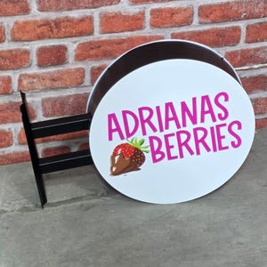 May include: A round, white sign with the text "ADRIANAS BERRIES" in pink, featuring a graphic of a chocolate-covered strawberry. The sign is mounted on a black metal bracket against a brick wall.