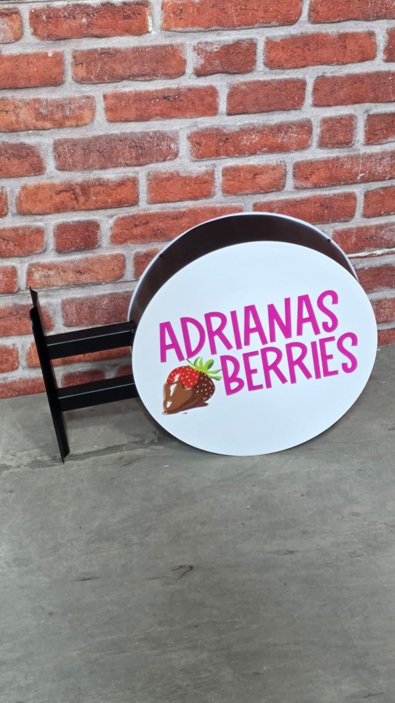 May include: A round, white sign with the text "ADRIANAS BERRIES" in pink, featuring a graphic of a chocolate-covered strawberry. The sign is mounted on a black metal bracket against a brick wall.