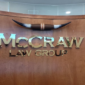 May include: A wall-mounted sign featuring the text "McCRAW LAW GROUP" in polished gold-toned metal letters. Above the text is a decorative longhorn steer horn mount. To the left, a vertical sword-shaped emblem is integrated into the wood paneling.