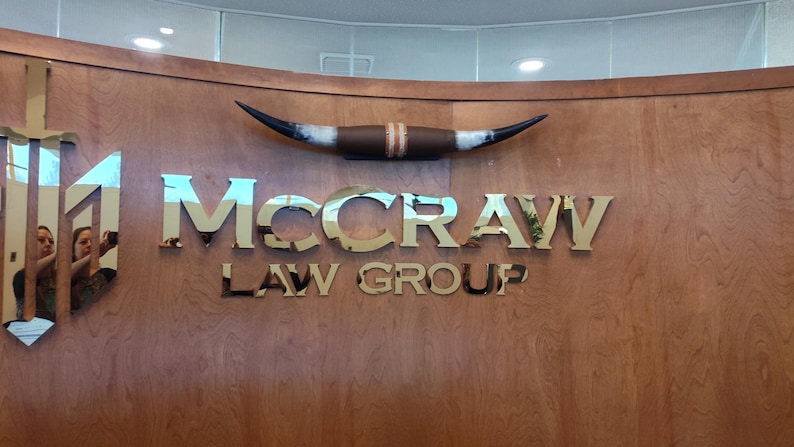 May include: A wall-mounted sign featuring the text "McCRAW LAW GROUP" in polished gold-toned metal letters. Above the text is a decorative longhorn steer horn mount. To the left, a vertical sword-shaped emblem is integrated into the wood paneling.