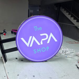 May include: A round, illuminated sign for "The VAPA SHOP" in purple and white. The sign is mounted on a black bracket and sits on a work table. The shop name is displayed in a modern font.