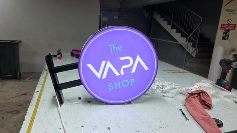 May include: A round, illuminated sign for "The VAPA SHOP" in purple and white. The sign is mounted on a black bracket and sits on a work table. The shop name is displayed in a modern font.